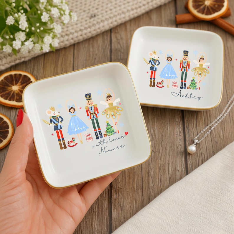 May include: Two square, white ceramic trinket dishes with gold rims. Each dish features a colorful illustration of a Nutcracker-themed design with a mouse, ballerina, and toy soldier. One dish has the name "Ashley" and the other says "with love Nannie."
