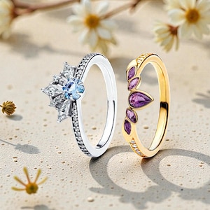 May include: Two rings are displayed. One is silver with a snowflake design and a light blue center stone. The other is gold with purple teardrop-shaped stones. The rings are on a light-colored surface with flowers in the background.
