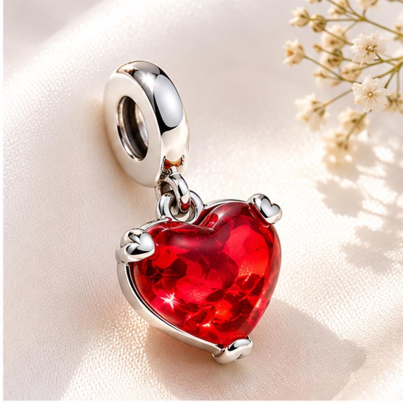 Op de afbeelding: Een zilveren bedel met een rode hartvormige hanger. Het hart heeft een facetachtig uiterlijk en is versierd met kleine zilveren hartjes. De bedel heeft een lus om aan een armband of ketting te bevestigen.