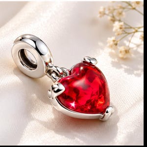 Op de afbeelding: Een zilveren bedel met een rode hartvormige hanger. Het hart is helderrood met een facetachtig uiterlijk en de bedel heeft een lus om aan een armband of ketting te bevestigen. De bedel is gemaakt van zilver.