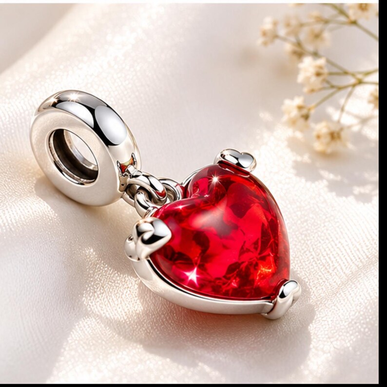 Op de afbeelding: Een zilveren bedel met een rode hartvormige hanger. Het hart is helderrood met een facetachtig uiterlijk en de bedel heeft een lus om aan een armband of ketting te bevestigen. De bedel is gemaakt van zilver.