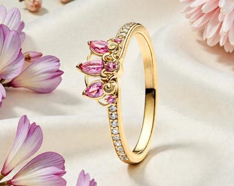May include: A gold-colored ring with a crown design featuring pink gemstones. The ring has a band lined with small, clear stones. The background includes pink flowers and a white, silky fabric.