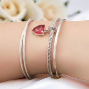 Op de afbeelding: Een zilveren armbandenset met drie armbanden. E&eacute;n armband heeft een rode hartvormige bedel. De andere twee armbanden zijn eenvoudige zilveren banden. De armbanden worden om een pols gedragen.