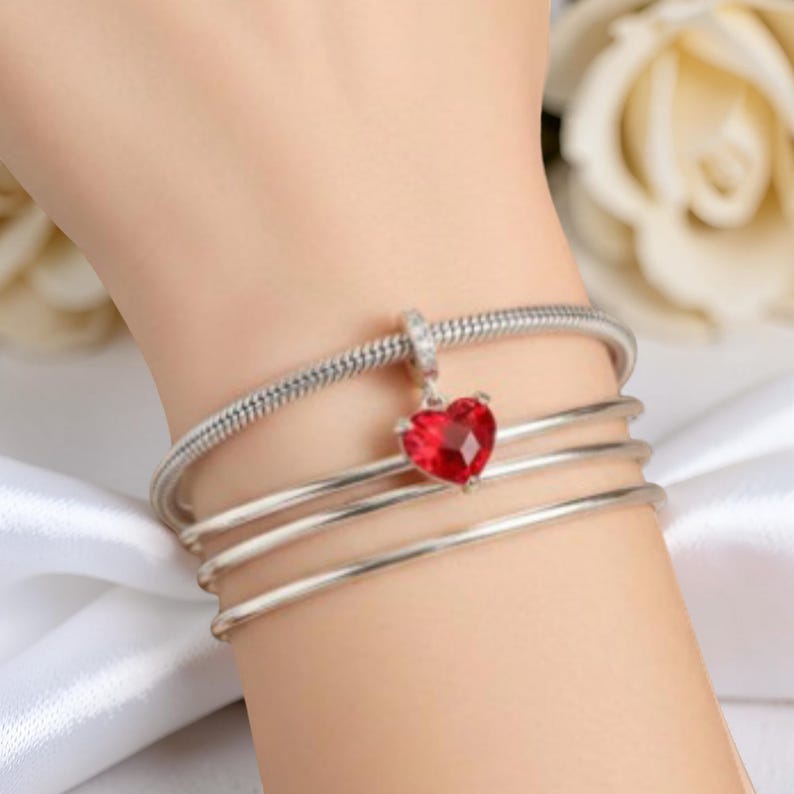 Op de afbeelding: Een zilveren armband met een rode hartvormige bedel. De bedel is een gefacetteerde, robijnrode edelsteen. De armband heeft een slangenkettingontwerp en wordt om de pols gedragen.
