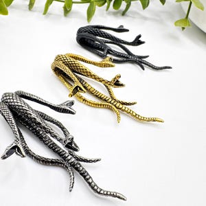 May include: Three metal snake hair clips in black, gold, and silver. Each clip features a detailed snake design with multiple heads and a textured body. The clips are arranged on a white surface, with green foliage in the background.
