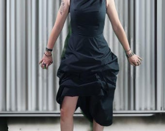 May include: A dark navy blue dress with a fitted bodice and a draped, asymmetrical skirt. The dress is paired with black ankle boots. The background features a textured, vertical-striped wall.