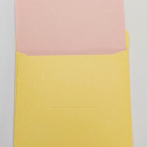 May include: A yellow envelope with a pink card inside. The envelope has a small, triangular flap and a horizontal indentation. The card is visible above the envelope's opening.
