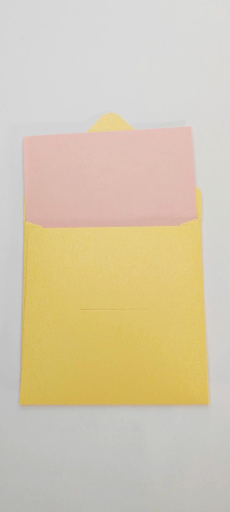 May include: A yellow envelope with a pink card inside. The envelope has a small, triangular flap and a horizontal indentation. The card is visible above the envelope's opening.