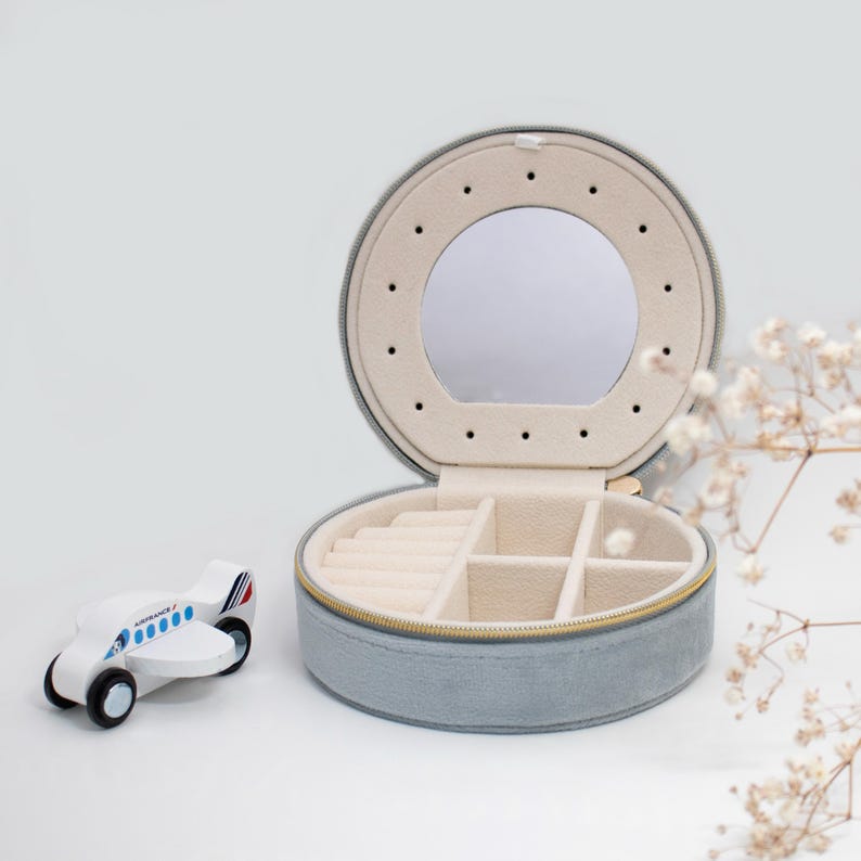 Puede incluir: Una caja de joyer&iacute;a redonda, azul claro, con espejo y cremallera dorada. El interior es beige con ranuras para anillos y compartimentos. Un avi&oacute;n de juguete de madera con detalles azules y rojos est&aacute; al lado.