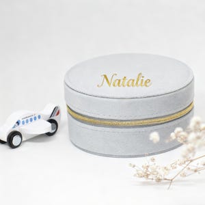 Puede incluir: Una caja de joyer&iacute;a redonda gris claro con cremallera dorada y el nombre "Natalie" en letras doradas. Un avi&oacute;n de juguete de madera blanca con detalles azules est&aacute; a la izquierda. La caja tiene unos 10 cm de di&aacute;metro.