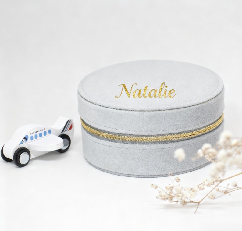 Puede incluir: Una caja de joyer&iacute;a redonda gris claro con cremallera dorada y el nombre "Natalie" en letras doradas. Un avi&oacute;n de juguete de madera blanca con detalles azules est&aacute; a la izquierda. La caja tiene unos 10 cm de di&aacute;metro.