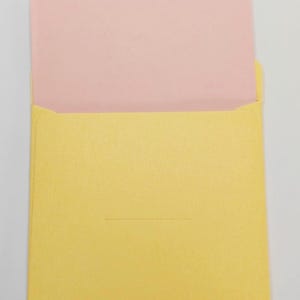 Puede incluir: Un sobre amarillo con una tarjeta rosa en su interior. El sobre tiene una peque&ntilde;a solapa triangular y una l&iacute;nea horizontal cerca de la parte inferior. El fondo es blanco liso.