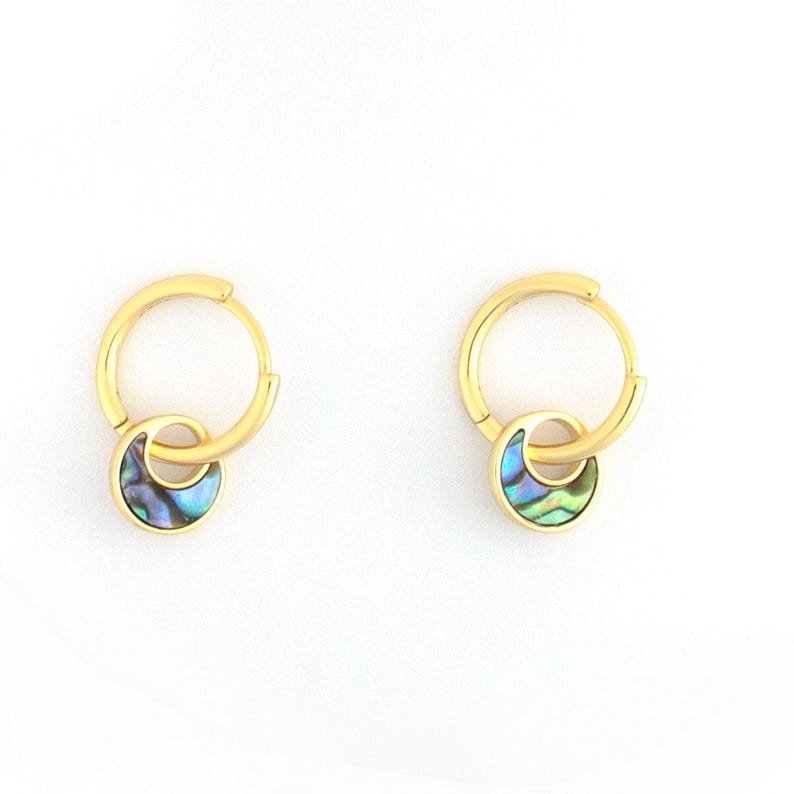 May include: A pair of gold-colored hoop earrings. Each earring features a small, iridescent, abalone shell charm in shades of blue, green, and purple. The earrings are displayed against a white background.