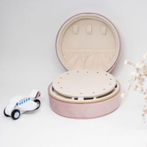 Puede incluir: Una caja de joyer&iacute;a redonda de color rosa p&aacute;lido con un interior crema y una bandeja extra&iacute;ble. La caja est&aacute; abierta, revelando soportes para aretes y un peque&ntilde;o compartimento. Un peque&ntilde;o avi&oacute;n de juguete de madera est&aacute; a la izquierda de la caja.