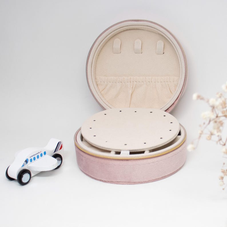 Puede incluir: Una caja de joyer&iacute;a redonda de color rosa p&aacute;lido con un interior crema y una bandeja extra&iacute;ble. La caja est&aacute; abierta, revelando soportes para aretes y un peque&ntilde;o compartimento. Un peque&ntilde;o avi&oacute;n de juguete de madera est&aacute; a la izquierda de la caja.