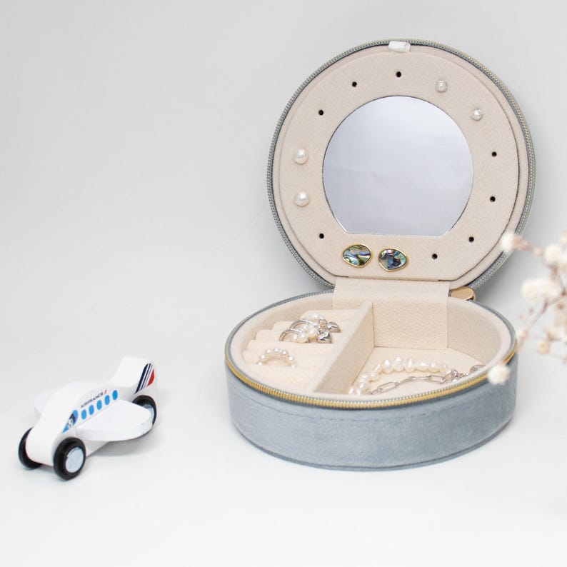 Puede incluir: Una caja de joyer&iacute;a redonda, azul claro, con espejo y detalles de perlas. La caja abierta revela compartimentos con anillos y un collar de perlas. A la izquierda hay un peque&ntilde;o avi&oacute;n de juguete de madera.