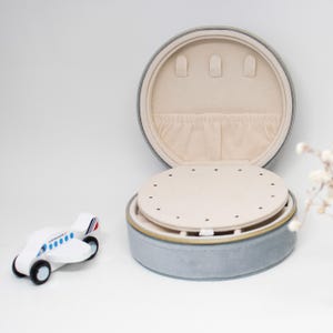 Puede incluir: Caja de joyer&iacute;a redonda gris abierta con interior beige y bandeja extra&iacute;ble. La caja tiene una cremallera dorada. Hay un peque&ntilde;o avi&oacute;n de juguete de madera al lado. La caja est&aacute; dise&ntilde;ada para guardar collares, pendientes y anillos.
