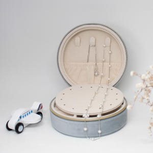 Puede incluir: Una caja de joyer&iacute;a redonda azul claro con interior beige, abierta para exhibir collares. Se ven un collar de plata con colgante de perla y un collar de perlas. Un avi&oacute;n de juguete de madera est&aacute; a la izquierda.