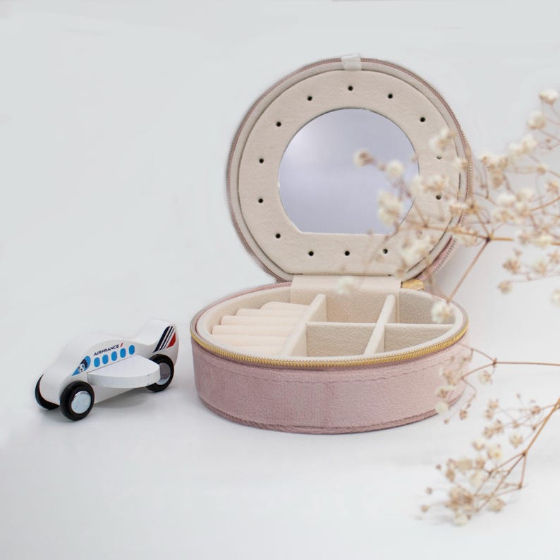 Puede incluir: Una caja de joyer&iacute;a redonda de color rosa empolvado con un espejo y compartimentos para guardar. Un peque&ntilde;o avi&oacute;n de juguete de madera con el texto "Air France" est&aacute; al lado de la caja. La caja de joyer&iacute;a est&aacute; abierta, revelando un interior beige.