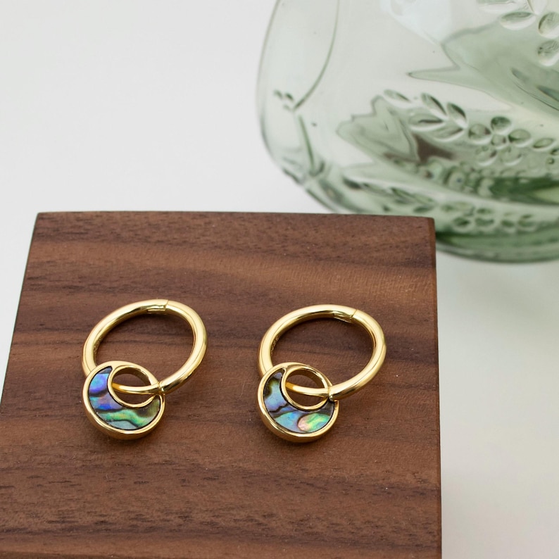 May include: A pair of gold-colored hoop earrings. Each earring features a circular design with a smaller, iridescent abalone shell accent. The earrings are displayed on a wooden surface.