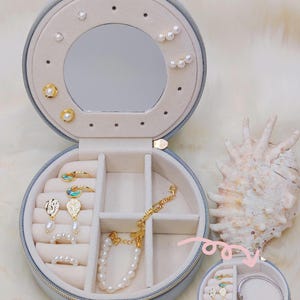 Puede incluir: Una caja de joyer&iacute;a redonda azul claro, abierta para mostrar varias joyas. Incluye pendientes, anillos y pulseras con perlas y detalles dorados. Un espejo est&aacute; dentro. Una concha marina est&aacute; al lado.
