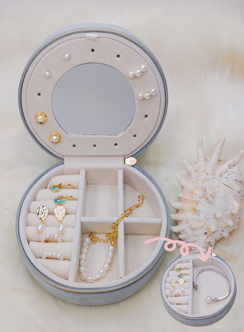 Puede incluir: Una caja de joyer&iacute;a redonda azul claro, abierta para mostrar varias joyas. Incluye pendientes, anillos y pulseras con perlas y detalles dorados. Un espejo est&aacute; dentro. Una concha marina est&aacute; al lado.