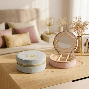 May include: Two jewellery boxes on a wooden surface. One is a round, light blue box with "Natalie" in gold script. The other is a pink, open, round box with a mirror and compartments for jewellery. A necklace and pearls are also visible.