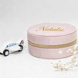 Puede incluir: Una caja de joyer&iacute;a redonda de color rosa claro con el nombre "Natalie" en letras doradas. La caja tiene una cremallera dorada y est&aacute; junto a un avi&oacute;n de juguete de madera blanco con detalles azules y rojos.