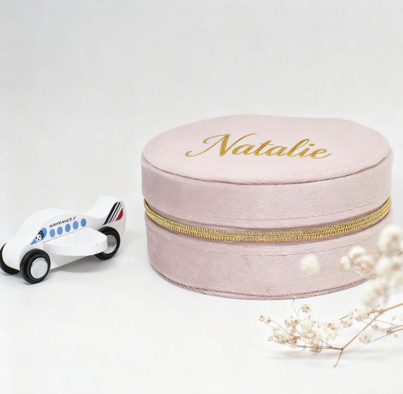 Puede incluir: Una caja de joyer&iacute;a redonda de color rosa claro con el nombre "Natalie" en letras doradas. La caja tiene una cremallera dorada y est&aacute; junto a un avi&oacute;n de juguete de madera blanco con detalles azules y rojos.
