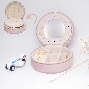 Puede incluir: Una caja de joyer&iacute;a redonda rosa con espejo y compartimentos para aretes, anillos y collares. La caja est&aacute; abierta, mostrando varias joyas de oro y perlas. Un peque&ntilde;o avi&oacute;n de madera est&aacute; al lado.