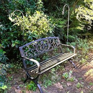 May include: A black metal garden bench with ornate scrollwork on the backrest and curved armrests. The bench is set in a garden with lush green foliage and two white metal plant hangers.