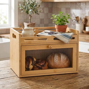 May include: A rectangular bamboo bread box with a drop-down door featuring a ribbed glass panel. The top surface holds a ceramic jar with a wooden spoon, a small potted green plant, and a folded white cloth with blue stripes and dried lavender.