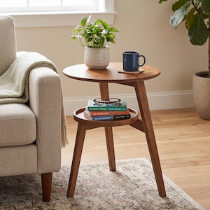 May include: A round wooden side table with three angled legs and a lower circular shelf. The top surface holds a green potted plant in a textured white pot and a dark blue mug. The lower shelf holds three stacked books and eyeglasses.