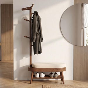 May include: A dark wood coat rack with a deer-shaped top hook and three additional side hooks. A dark gray trench coat hangs from the rack. Below is a bench with a cream-colored cushion and a lower shelf holding two pairs of shoes.