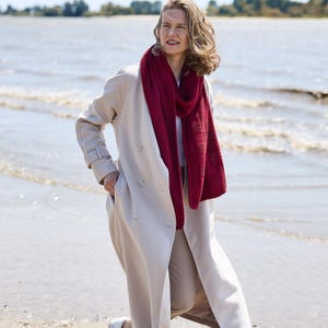 May include: A woman wearing a light beige trench coat and a deep red scarf. The scarf is draped around the neck and shoulders. The woman is standing on a beach with the sea in the background.