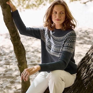 Pode incluir: Uma mulher vestindo um suéter azul marinho com um padrão geométrico branco no peito e ombros. O suéter tem mangas compridas e gola redonda. Ela está usando calças brancas e sentada em um galho de árvore.