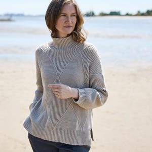 May include: A light beige turtleneck sweater with a geometric diamond pattern on the front. The sweater has long sleeves and a ribbed hem. The model is wearing the sweater on a beach.