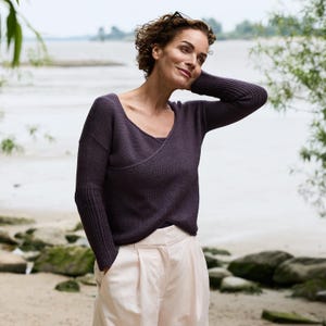 Könnte beinhalten: Eine Frau trägt einen dunkelvioletten Pullover im Wickelstil mit langen Ärmeln und eine hellrosa Hose. Der Hintergrund zeigt einen Strand und Wasser.