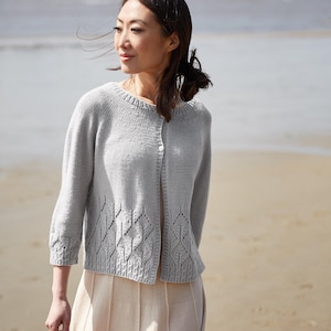 Könnte beinhalten: Hellgraue Strickjacke mit Knöpfen, rundem Ausschnitt und Dreiviertelärmeln. Der Strickpullover hat ein dekoratives Blättermuster am unteren Rand. Die Frau trägt einen beigen Rock und steht am Strand.