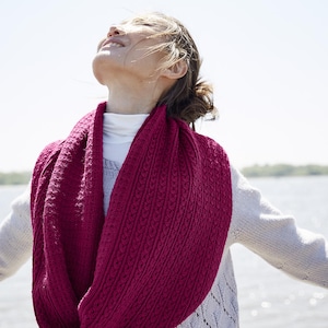 Könnte beinhalten: Ein gestrickter Rundschal in tiefem Magenta. Der Schal hat ein strukturiertes Zopfmuster und wird über einem weißen Rollkragenpullover getragen. Der Hintergrund zeigt einen hellen Himmel und Wasser. Der Schal ist ca. 150 cm lang.