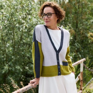 May include: A color-blocked cardigan in gray, olive green, and navy blue. The V-neck sweater has a button closure and a pocket. The sleeves and lower hem are olive green, with navy blue accents on the sleeves and neckline. The woman is wearing glasses and a white dress.