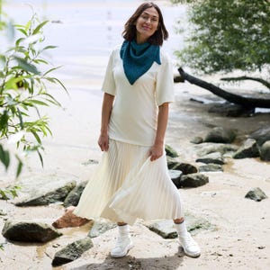 May include: A woman wearing a teal knitted shawl, a cream-colored t-shirt, and a pleated skirt. She is also wearing white sneakers and socks. The background is a beach scene with rocks and water.