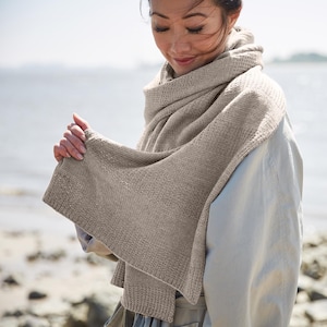 May include: A beige knitted scarf draped around the shoulders. The scarf has a textured pattern and is wrapped around the neck and shoulders. The person is wearing a light-colored coat. The background is a beach with water and sky.