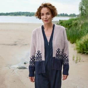 May include: A light pink cardigan with a navy blue patterned design on the lower section and sleeves. The cardigan is worn over a navy blue jumpsuit. The background shows a beach and water.
