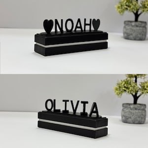 Op de afbeelding: Zwart gepersonaliseerd naamplaatje met de naam "NOAH" en hartsymbolen aan weerszijden. De naam is in een vetgedrukte, schreefloze lettertype. Het naamplaatje staat op een rechthoekige zwarte basis met een witte accentstreep. Een tweede naamplaatje met de naam "OLIVIA" wordt ook getoond.