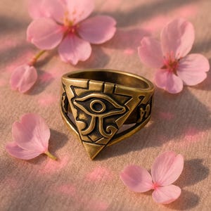 Pode incluir: Um anel de cor bronze com o símbolo do Olho de Hórus, rodeado por pétalas de flores de cerejeira rosa. O anel apresenta um design triangular com detalhes gravados e uma banda com padrões. O design lembra a arte egípcia antiga.