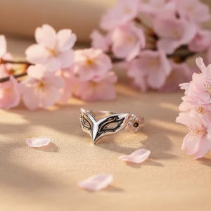 Op de afbeelding: Een zilveren ring met een maskerontwerp, met een wit en zwart kleurenschema. De ring staat tegen een achtergrond van roze kersenbloesems en bloemblaadjes, wat een delicate en artistieke compositie creëert.