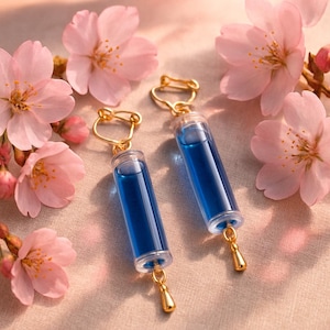 Puede incluir: Un par de pendientes con viales cilíndricos transparentes llenos de líquido azul, suspendidos de ganchos dorados. Cada pendiente tiene un pequeño dije dorado en forma de lágrima. Flores de cerezo rosas rodean los pendientes.