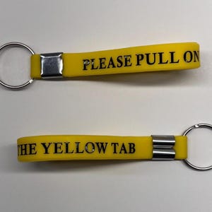 Puede incluir: Dos llaveros amarillos con anillas plateadas. Los llaveros tienen el texto "PLEASE PULL ON" y "THE YELLOW TAB" impreso en negro. Los llaveros están sobre una superficie blanca.