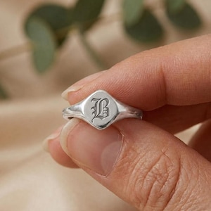 Puede incluir: Un anillo de sello de plata con acabado pulido y cara en forma de diamante. El centro del anillo está grabado con la letra B en una fuente gótica decorativa. La banda es lisa y se estrecha hacia la parte posterior del anillo.